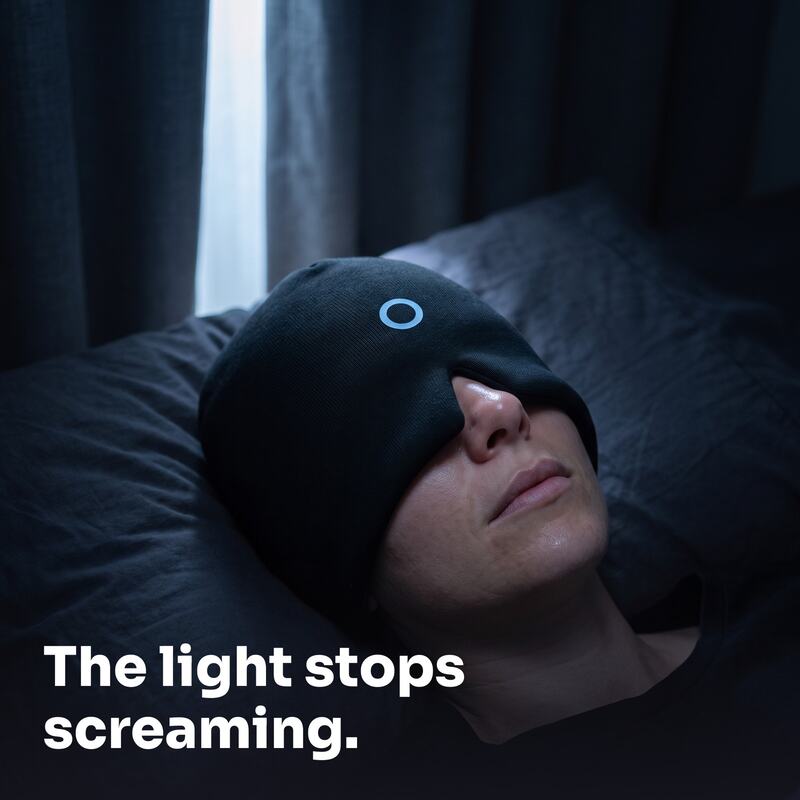 Woman lying in a dark bedroom wearing the DUSKHALO cap pulled down over her eyes
